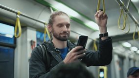 Handsome Caucasian man with long hair tied in bun standing in tram holding strap and smiling at camera. Male wearing dark jacket using smartphone with one hand during evening ride on public transport. - Powered by Shutterstock - Get 15% off with code: PIKWIZARD15