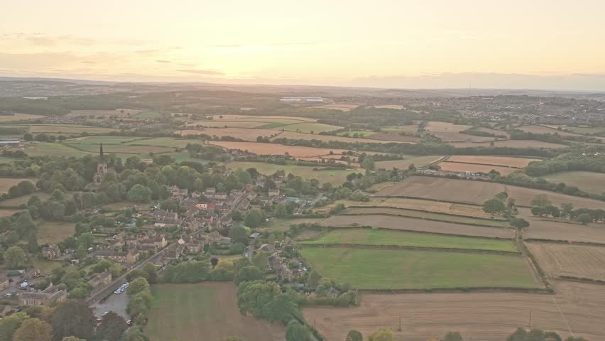Drone pulls out over Wentworth into the landscape with its Gothic bell tower of the Holy Trinity Church, clustered rooftops, and tree-lined fields fading into rolling hills under a glowing sunset sky.