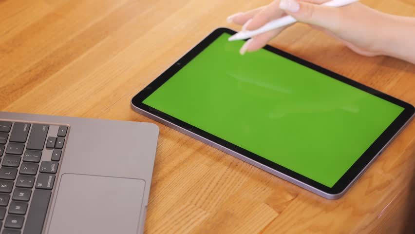 Person is using a tablet with a green screen. A laptop is also on the table. The tablet is on the right side of the laptop