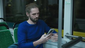 Caucasian man with beard wearing blue sweater sitting by window holding smartphone. Male scrolling social media while waiting for bus stop during night ride. Relaxed moment on public transport. - Powered by Shutterstock - Get 15% off with code: PIKWIZARD15