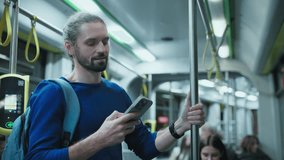 Caucasian man with beard wearing blue sweater standing on city bus holding pole with one hand and smartphone in other. Male reading online news during evening commute home from work. - Powered by Shutterstock - Get 15% off with code: PIKWIZARD15