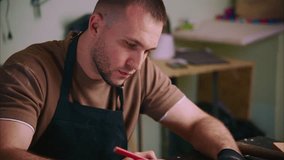 Skilled craftsman wearing protective black gloves and leather apron carefully drawing woodworking project design on paper while sitting at wooden workbench inside professional carpentry workshop - Powered by Shutterstock - Get 15% off with code: PIKWIZARD15