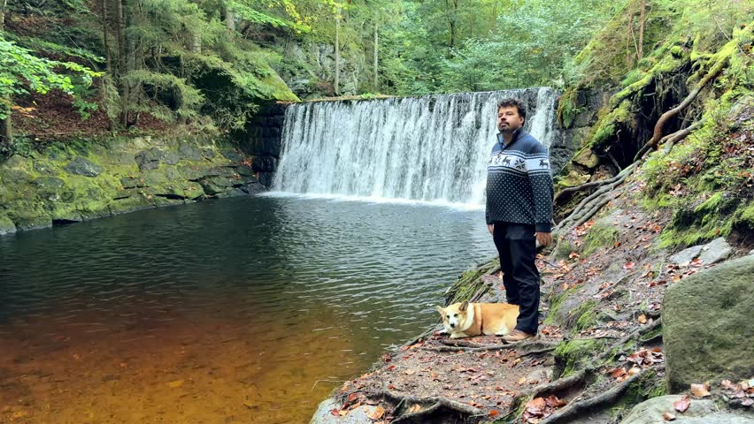Man stands by serene waterfall with dog, camera slowly zooms in on tranquil scene