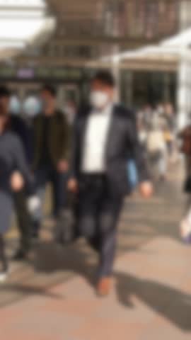Slow motion vertical clip of Japanese pedestrians walking on tiled city square. Framing is defocused to emphasize atmosphere, rhythm, and social repetition.