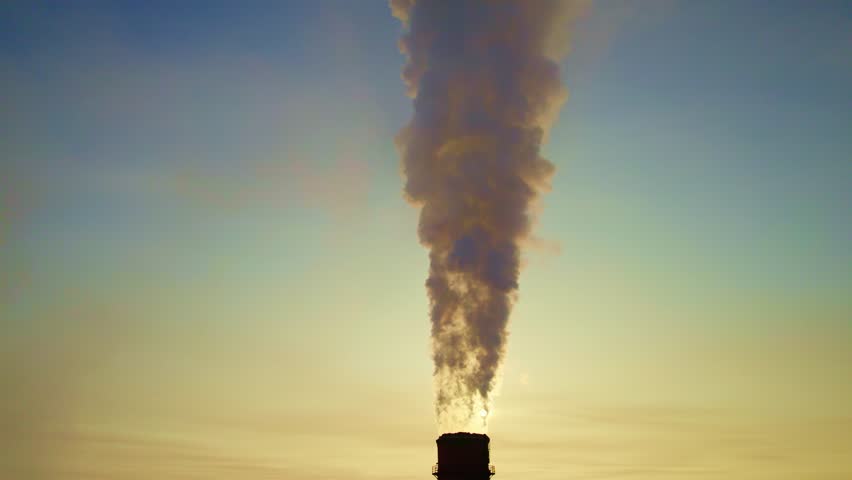Thick smoke rises from a smokestack as the sun sets in the background, casting a dramatic silhouette against the evening sky. The scene highlights industrial activity in an urban setting. - Powered by Shutterstock - Get 15% off with code: PIKWIZARD15