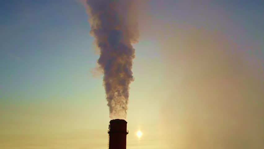 A towering industrial chimney emits thick smoke into the evening sky as the sun sinks below the horizon. The scene captures the stark contrast of pollution and natural beauty.