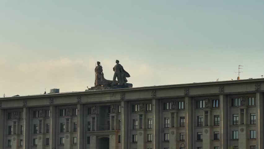 A horizontal, long-shot captures the dramatic silhouettes of large, socialist realist statues depicting a worker and a kolkhoz woman (collective farmer) on the rooftop of a monumental Soviet-era