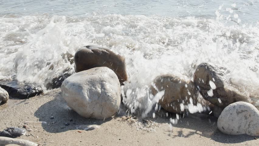 Powerful ocean waves crash over a cluster of large, smooth beach rocks on a sandy shore in bright sunlight. The water surges and retreats, demonstrating the relentless movement of the sea.