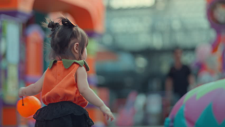 Cute little Asian girl wearing orange pumpkin costume holding jack o lantern bucket outdoors during Halloween festival, adorable child enjoying autumn holiday celebration with festive background.