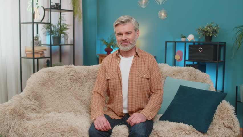 Mature smiling man sitting on home sofa waving his hand in greeting hello hi welcome. Middle-aged guy looks friendly and happy, expressing warmth and a welcoming attitude in a comfortable living room