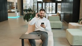 Happy relaxed Caucasian young businessman wearing headphones listening favorite energetic disco music on smartphone. Male entrepreneur in formal relaxing, taking a break sitting in modern office lobby - Powered by Shutterstock - Get 15% off with code: PIKWIZARD15