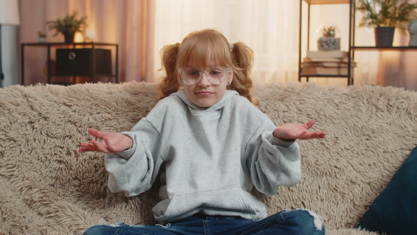 I dont know what to say. Young teenager girl shrugging shoulders with puzzled unsure expression at home. Caucasian child kid sitting on a sofa, showing confusion and uncertainty with relatable gesture