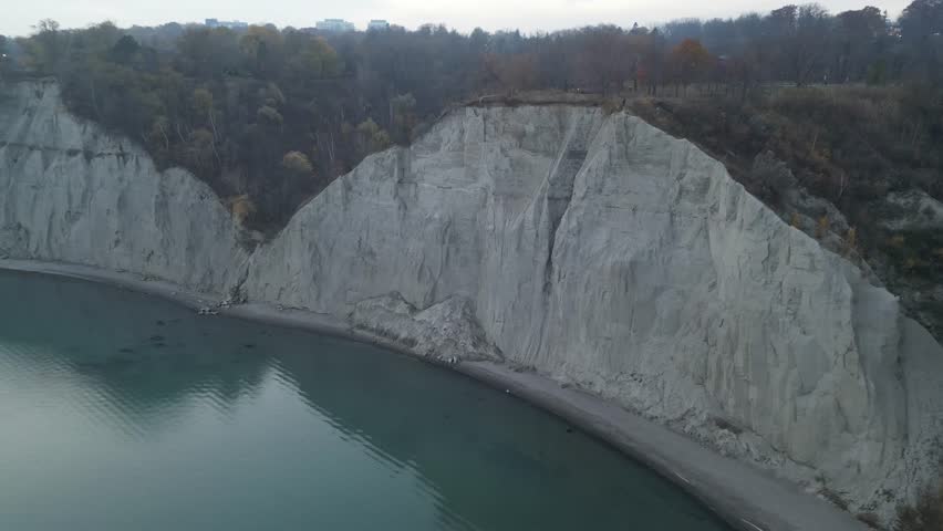 Aerial clip showing waves lapping at the base of the Bluffs, with green trees above and soft pastel sky. Ideal for relaxation, travel, or calm backdrop scenes