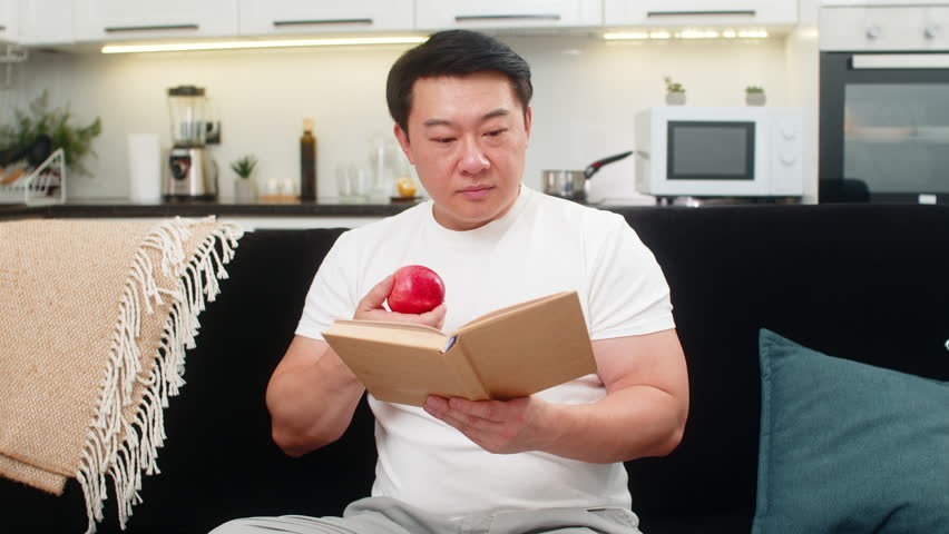 Asian man at home bites red apple enjoys juicy taste chews slow and smiles feeling fresh and awake. Chinese guy on sofa reads health tips in book marks page then eats more and feels fine and calm good