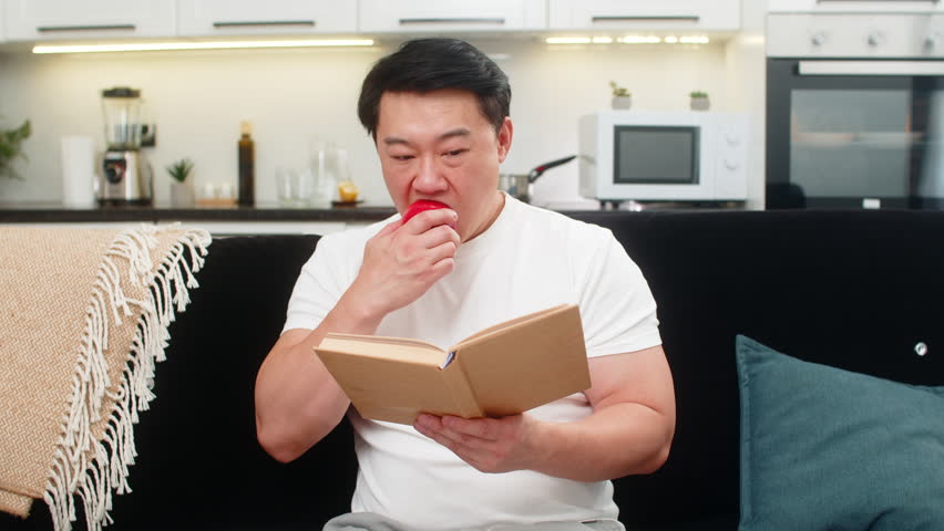 Asian man at home bites red apple enjoys juicy taste chews slow and smiles feeling fresh and awake. Chinese guy on sofa reads health tips in book marks page then eats more and feels fine and calm good