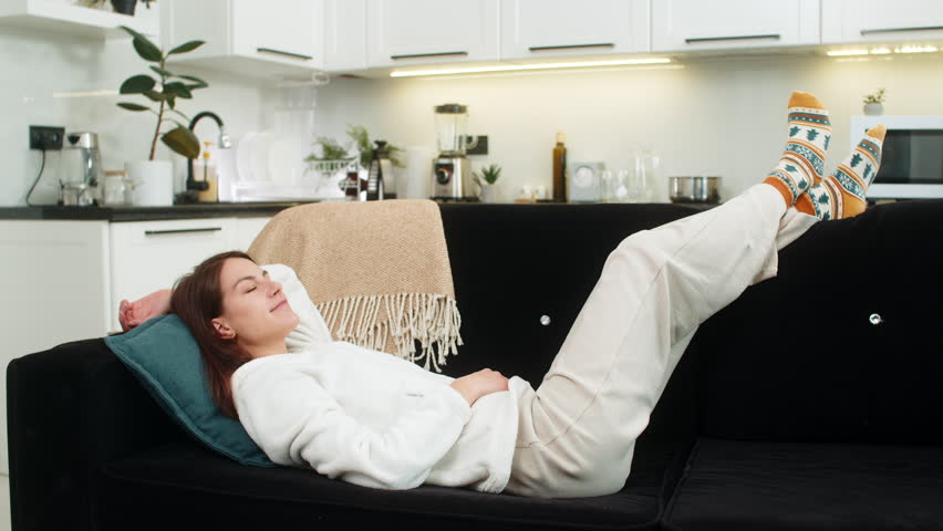 Young woman at home lies back and rests in cool air and calm hush from quiet home tech around. Caucasian girl on sofa lifts feet on back smiles enjoys stable climate control and low noise indoors here