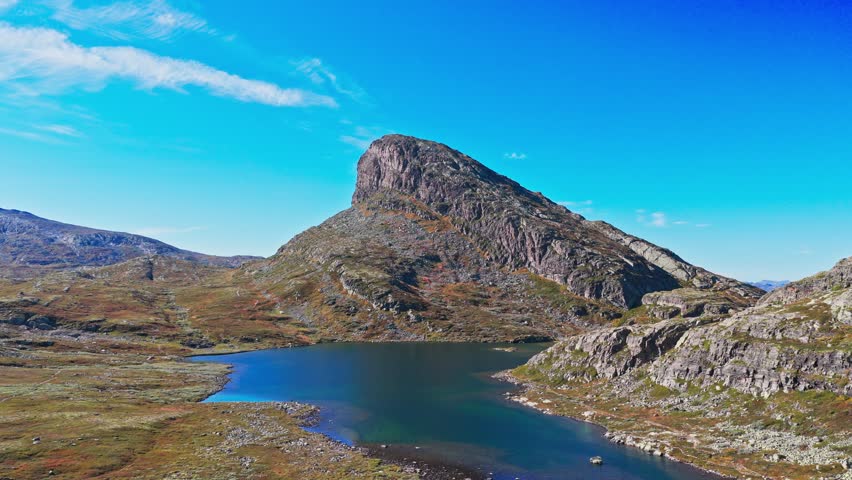 Breathtaking view of a majestic, conical mountain peak overlooking a serene, dark blue mountain lake under a bright sky in the rugged, autumnal Scandinavian wilderness.