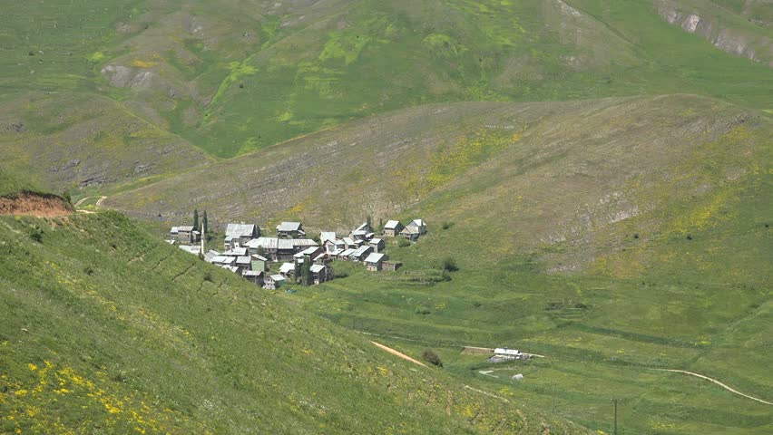 Panoramic view of a small village lying in the valley between tall mountains in Iran. Traditional houses and farmlands are surrounded by rugged slopes under warm daylight.