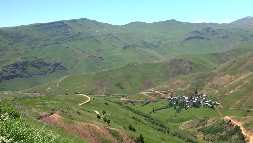 Panoramic view of a small village lying in the valley between tall mountains in Iran. Traditional houses and farmlands are surrounded by rugged slopes under warm daylight.
