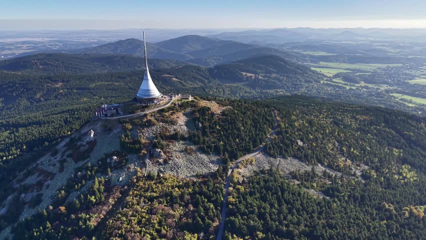 An aerial view shows a majestic tower on a mountain, surrounded by vibrant greenery and rolling hills