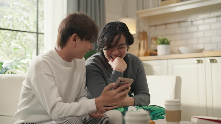 Asian LGBTQ couple shares intimate moments together on sofa, browsing cherished travel photos and precious memories on smartphone while enjoying quality weekend bonding time at comfortable home 