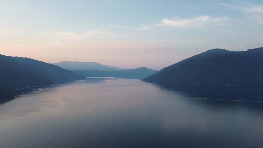 Breathtaking aerial view flying above a blanket of low fog on a vast mountain lake. The still water reflects the soft colors of the dawn sky, creating a serene and mystical scene.