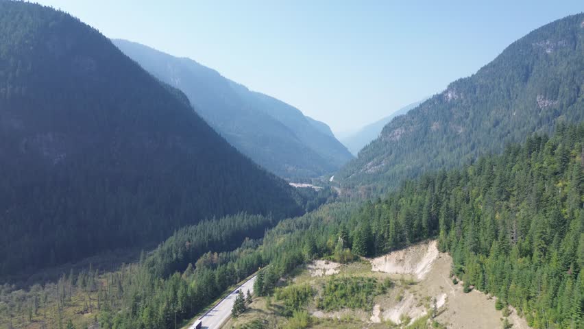 A stunning aerial view looking down a deep mountain valley. A highway snakes through the dense pine forest, surrounded by steep, rugged mountainsides in sun and shadow.