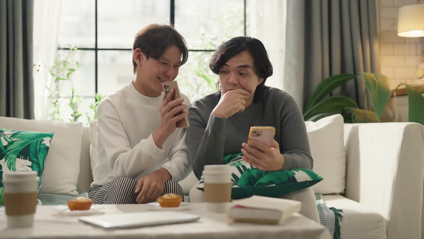 Asian LGBTQ couple shares intimate moments together on sofa, browsing cherished travel photos and precious memories on smartphone while enjoying quality weekend bonding time at comfortable home 