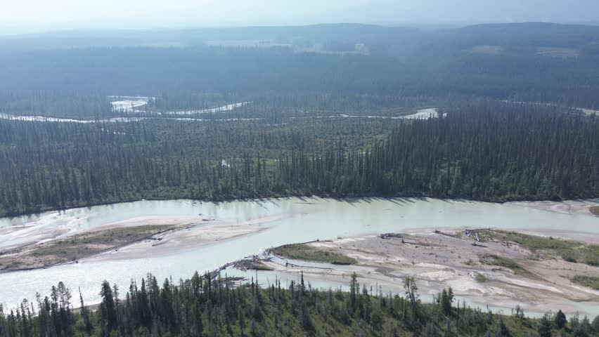 A sweeping aerial view of a massive, braided river carving through a dense forest. In the hazy distance, plumes of smoke rise from wildfires, showing nature's powerful cycle.