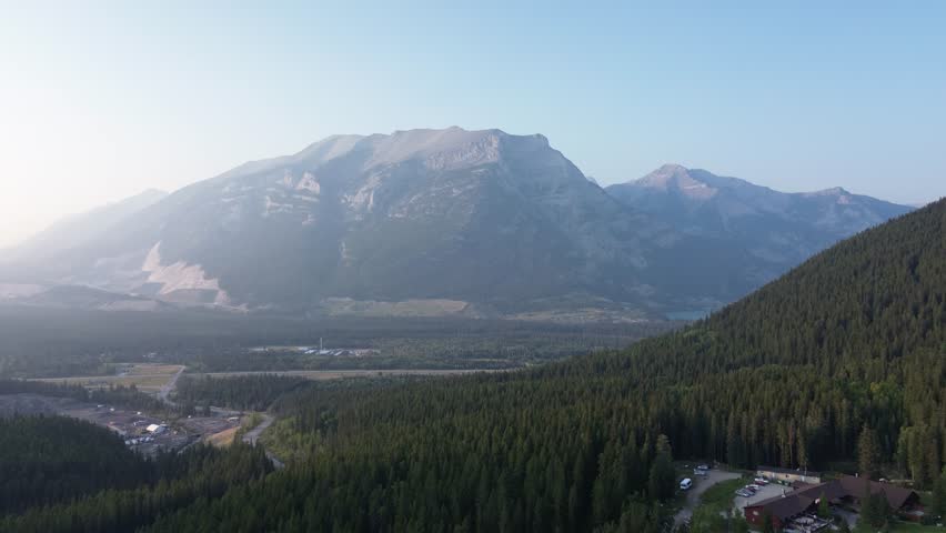 Stunning aerial footage shows an industrial complex in a remote mountain valley. Smoke rises from the site as the sun rises, casting a hazy glow over the epic mountain backdrop.
