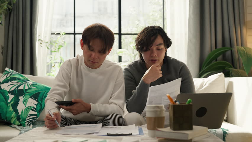 "Asian LGBTQ male couple reviews monthly financial bills and household expenses together at home, managing shared budget planning, savings goals and responsible money management for their future 