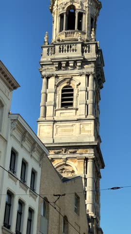 Close up of a magnificent, ornate Baroque church tower and belfry, built from light stone, reaching toward a clear blue sky, topped with a golden finial. Antwerp, Belgium