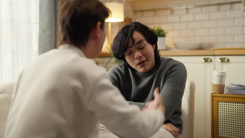 Asian gay male couple experiences relationship conflict during serious discussion on living room sofa at home daytime, working through disagreement and communication challenges to strengthen their par
