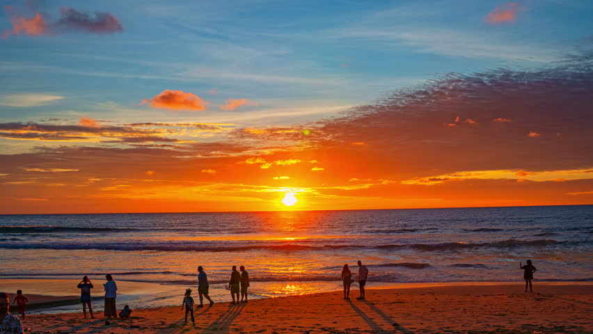 A stunning sunset over the ocean with people enjoying the sandy beach as the sky glows in shades of orange and blue. The warm light and tranquil waves create a peaceful and picturesque coastal scene.