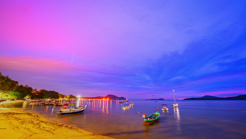 A peaceful twilight scene at Rawai Beach in Phuket. featuring fishing boats anchored along the calm shoreline under a soft purple and blue sky. reflections of lights shimmer on the tranquil water.