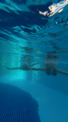 Two smiling girls swim underwater in a clear blue swimming pool, enjoying their time in a fun and carefree vacation moment.
