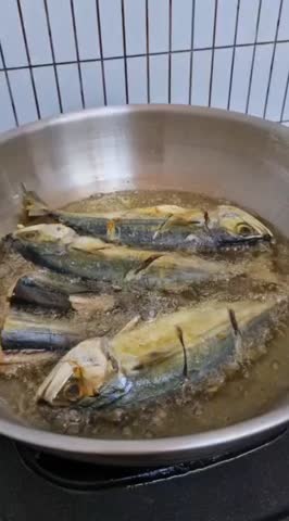 Several mackerels are deep-frying in a large wok with hot, sizzling oil in a kitchen setting.