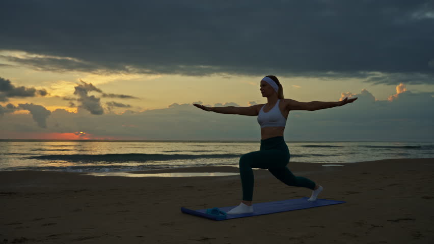 Fit woman striking warrior ii pose on yoga mat at sunrise on serene beach, woman outline accentuated by vibrant ocean horizon in background 