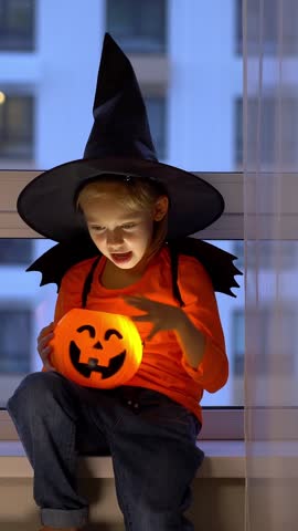 Girl sits by window in witch hat and conjures over halloween pumpkin lantern in evening. Child celebrates halloween. Vertical shot