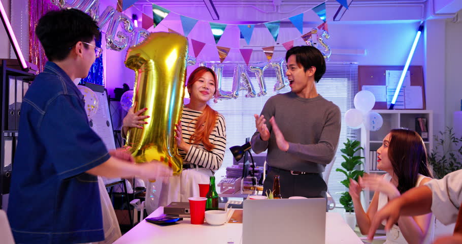 Group of young Asian coworkers man and woman celebrate their number one achievement at a fun office party. A modern business team enjoying success with balloons, cheers, and joyful expressions.