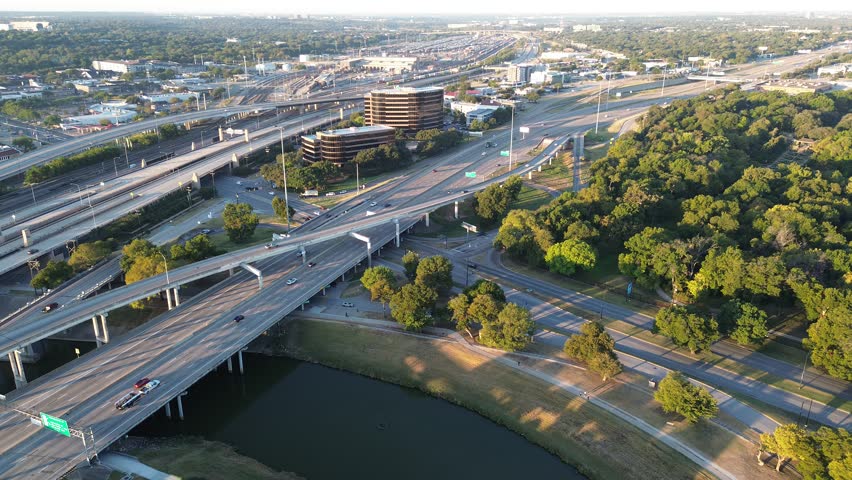 Lush urban park and tree canopy border the Trinity River, stacked highway ramps curving toward Fort Worth high-rise office buildings. Flyover blends mobility, green space, architectural density. USA
