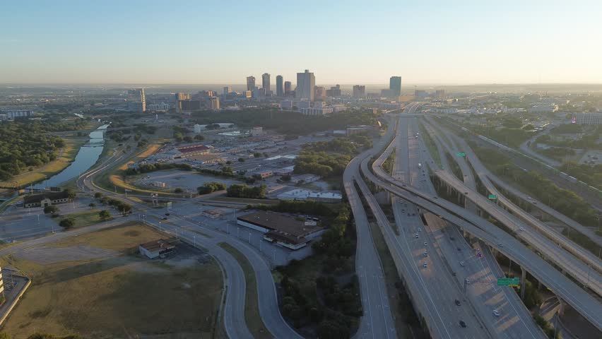 Curved Trinity River and stacked highway system lead into downtown Fort Worth, framed by light industrial buildings in foreground. Early morning light adds warmth to city layered commute corridor. USA