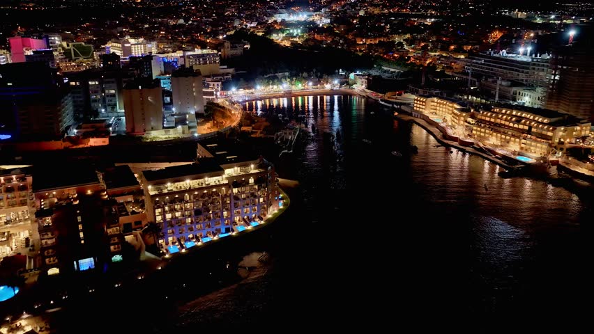 Aerial 4K view of St George Bay in Paceville’s nightlife hub: glowing rooftops, neon-lit towers and bustling streets pulse with energy in Malta’s entertainment capital