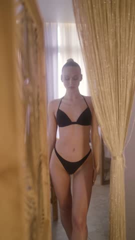 Gorgeous slender woman in black swimwear walking towards the camera and posing. Beautiful female model with perfect body in a luxurious interior