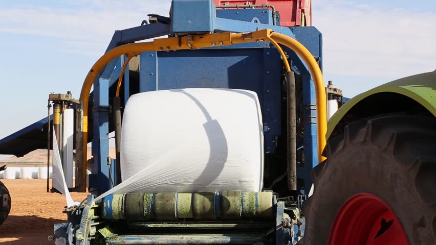 A bale wrapper is wrapping a bale of hay with plastic film in a field. The machine is blue and yellow. The bale is white. The field is brown. The sky is blue.