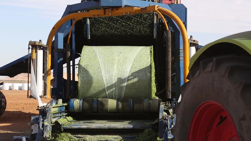 A bale wrapper is wrapping a bale of hay with plastic film in a field. The machine is blue and yellow. The bale is green. The field is brown. The sky is blue.