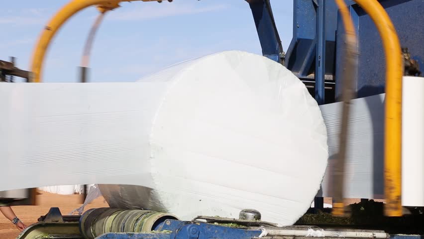 A bale wrapper is wrapping a bale of hay with plastic film in a field. The machine is blue and yellow. The bale is white. The field is brown. The sky is blue.