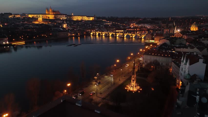 Aerial night view of Charles Bridge and Prague Castle above the Vltava River, with glowing city lights, reflections on water, and traffic moving through historic Prague