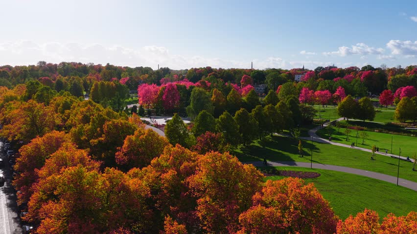 Aerial pan over a Riga park on a sunny autumn day, showing vibrant foliage, a ferris wheel, the National Library, and the old town skyline.