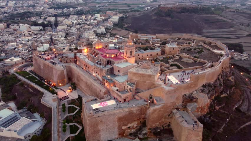 4K drone orbits the illuminated Citadella in Rabat, Gozo at dusk during the Light Festival: glowing ramparts, cathedral domes and festival colors create a cinematic cultural showcase ideal for travel